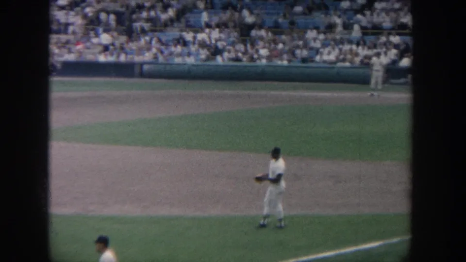 A 1964 New York baseball player stands in the field during game action, seen from a spectator's 8mm camera angle.