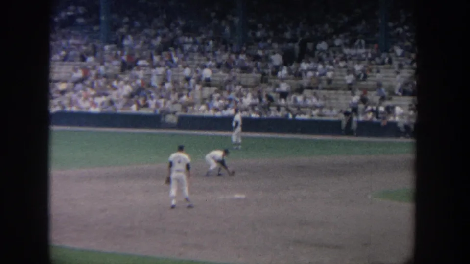 1964 New York baseball scene with an infielder fielding near a base while another player watches from the dirt and spectators sit beyond.