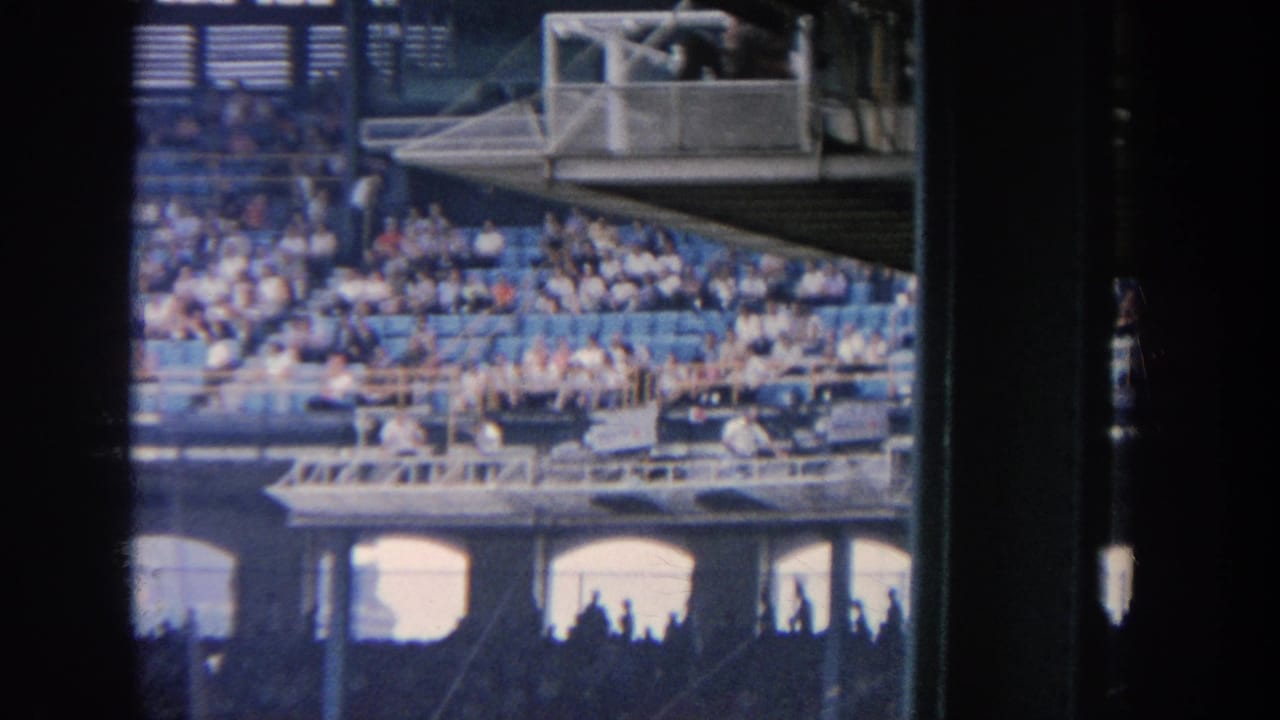 1964 New York baseball stadium view from the stands, showing spectators in blue seating, a press box and arched concourse openings through dark framing.