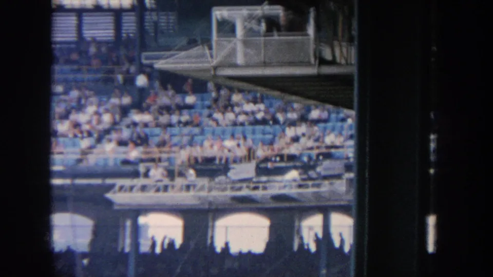 Home-movie view of a New York baseball stadium in 1964, showing spectators in tiered blue seats and a press-box area above arched stadium openings.