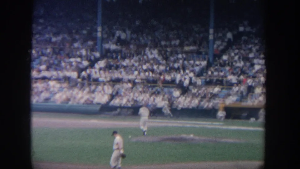 1964 New York baseball field view with a pitcher near the mound, another uniformed player on the grass and crowded stadium seating behind them.