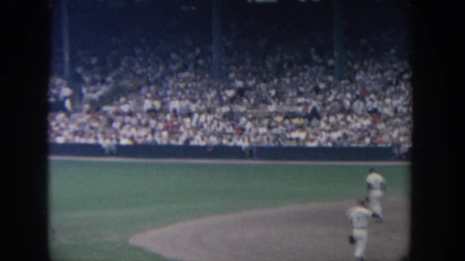1964 New York home-movie frame showing players near the infield edge and packed ballpark seating beyond the field.