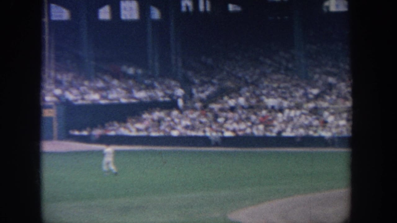 1964 New York 8mm frame showing a distant baseball player on the field, crowded stands, and dark foreground barriers.