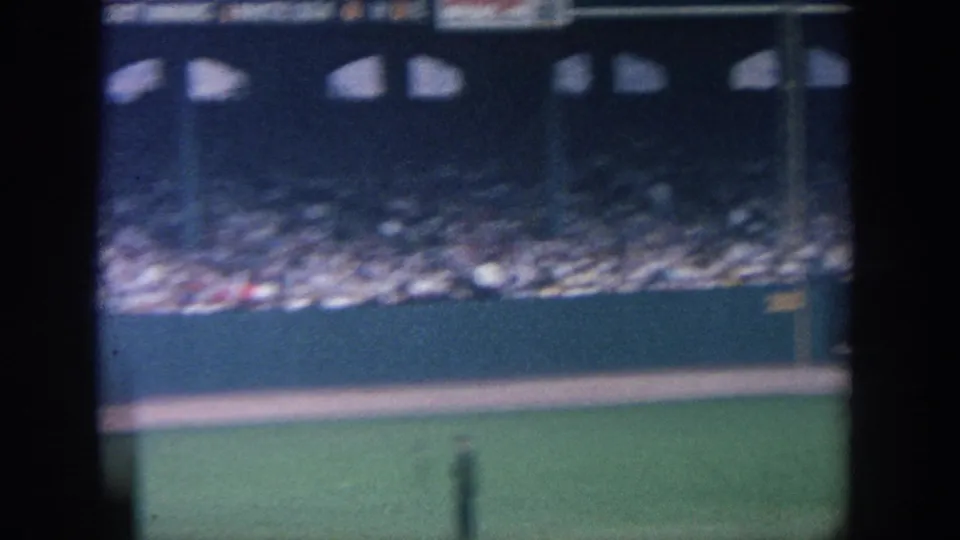 1964 New York home-movie view across a baseball stadium, with crowd seating, outfield wall, grass, and dark foreground posts.