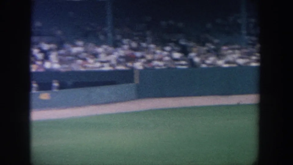 1964 New York stadium view showing the field edge, outfield wall, packed seating, and dark foreground obstruction in 8mm color.