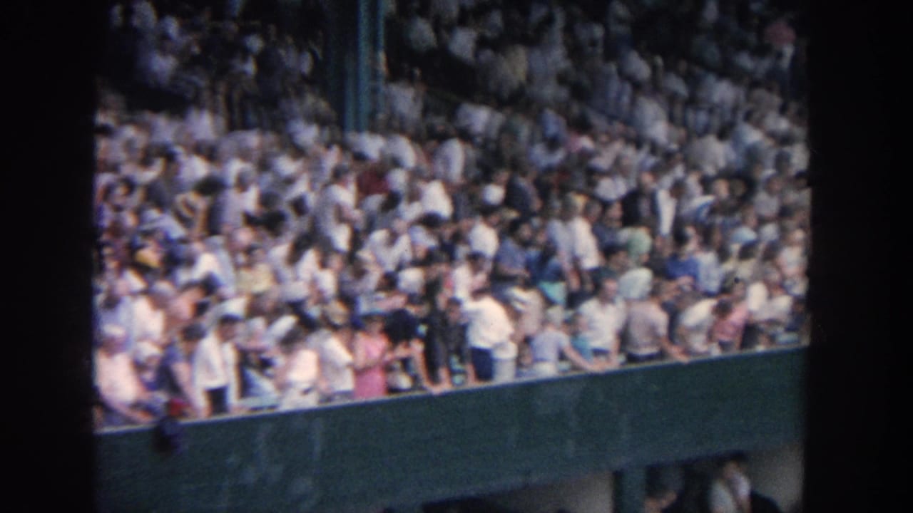 1964 New York home-movie view of packed baseball stands, with spectators behind a green stadium wall and dark 8mm frame edges.