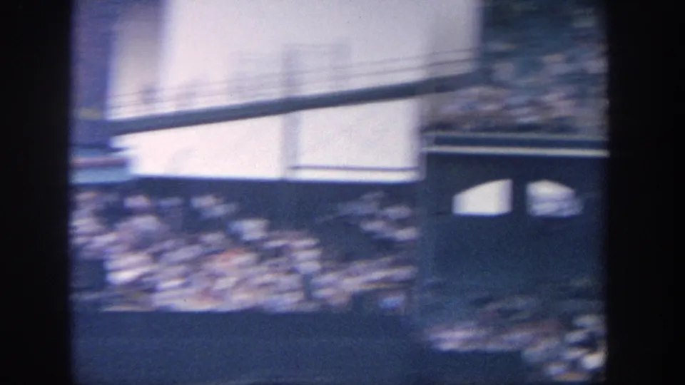 Handheld 8mm pan across a crowded baseball stadium, with seating and a green wall softened by motion blur and exposure shift.