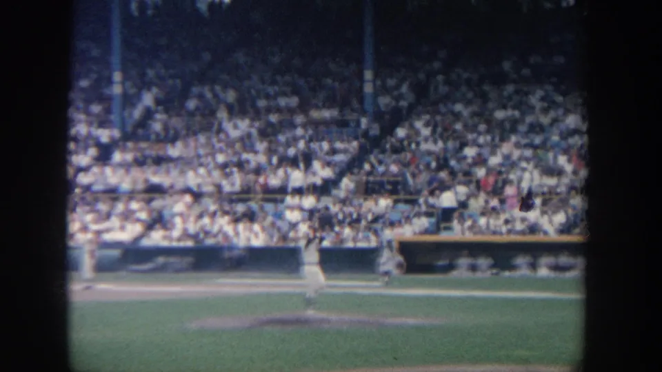 1964 New York home-movie frame showing a pitcher on the mound, field grass, dugout area and full stadium seating behind.