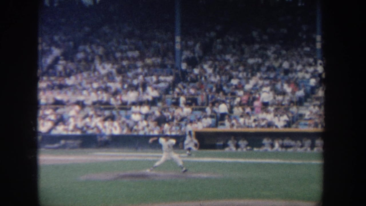 1964 New York 8mm frame showing a pitcher releasing the ball from the mound with packed stadium seats behind him.