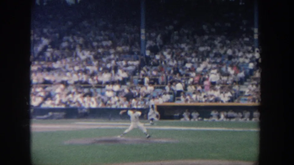 1964 New York 8mm frame showing a pitcher releasing the ball from the mound with packed stadium seats behind him.
