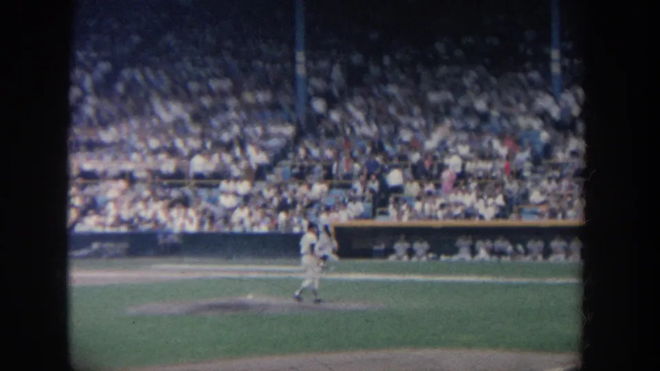 1964 New York 8mm frame with a baseball player on the field and a packed stadium crowd behind the dugout.