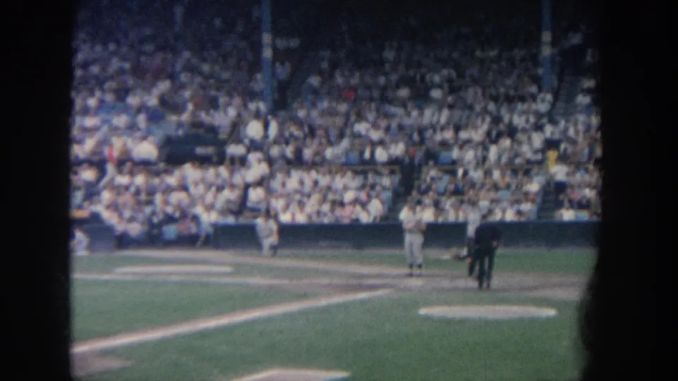 1964 New York 8mm frame showing the home plate area with batter, catcher, umpire, chalk lines, and spectators.