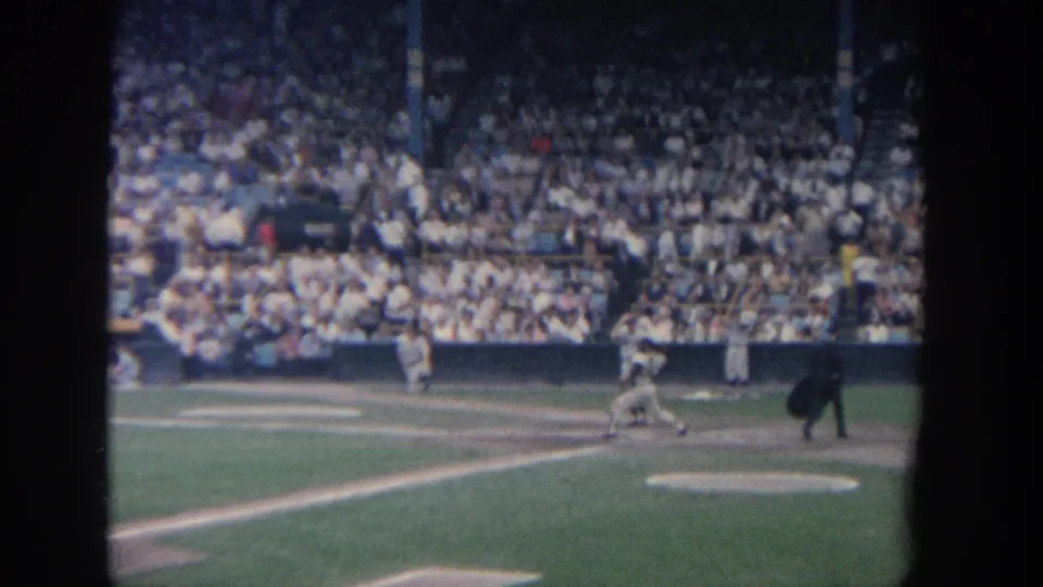1964 New York 8mm frame of a batter swinging near home plate with catcher, umpire, field markings, and grandstand crowd.
