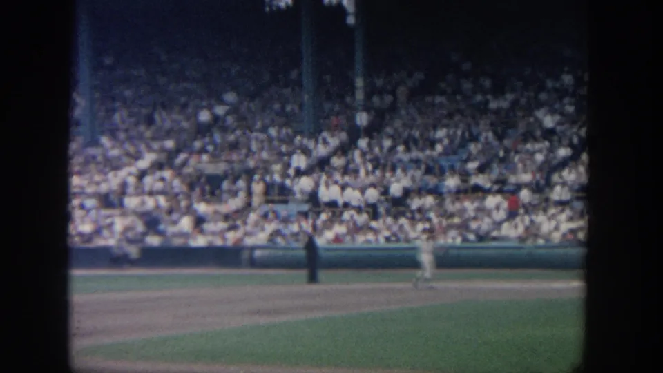 End-frame view of the baseball field with uniformed players, a dark-clad official-like figure, and packed stadium seating in 1964 New York footage.
