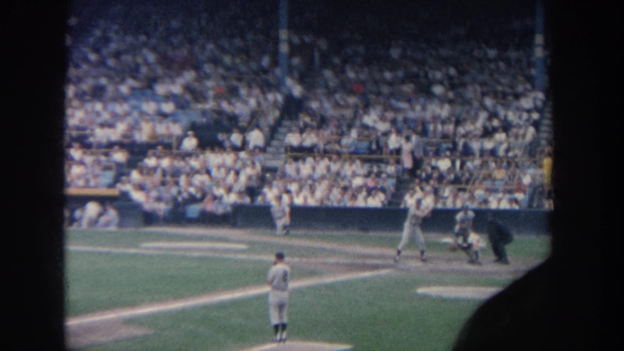 1964 New York 8mm frame showing a pitcher on the mound as a batter, catcher, and umpire wait at home plate before a full crowd.