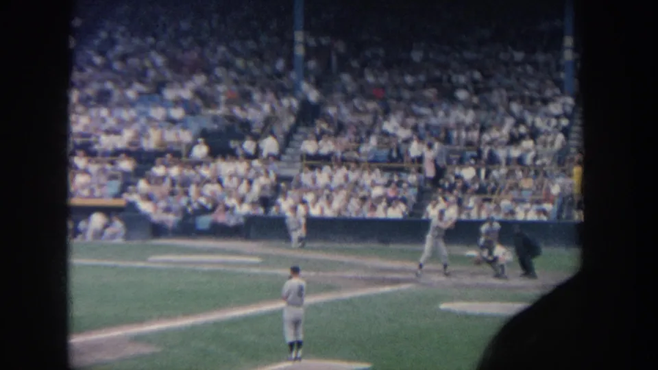 1964 New York 8mm frame showing a pitcher on the mound as a batter, catcher, and umpire wait at home plate before a full crowd.