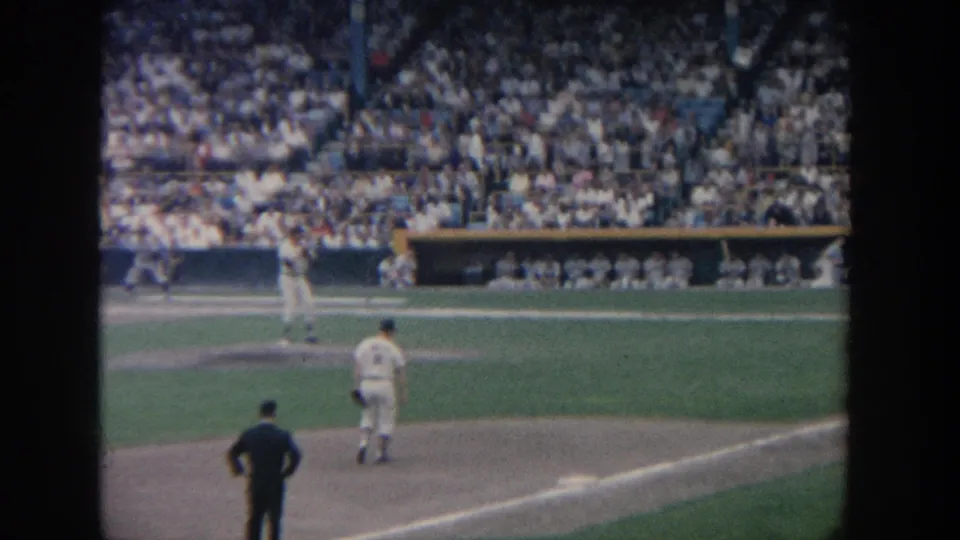 1964 New York 8mm frame showing infield players, an umpire, a dugout, and a packed baseball stadium crowd viewed from the stands.