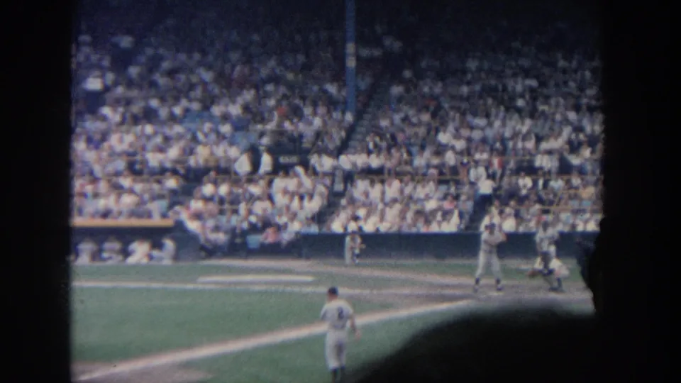 A 1964 New York professional baseball play unfolds near home plate, seen from a distant seat with a packed grandstand behind.