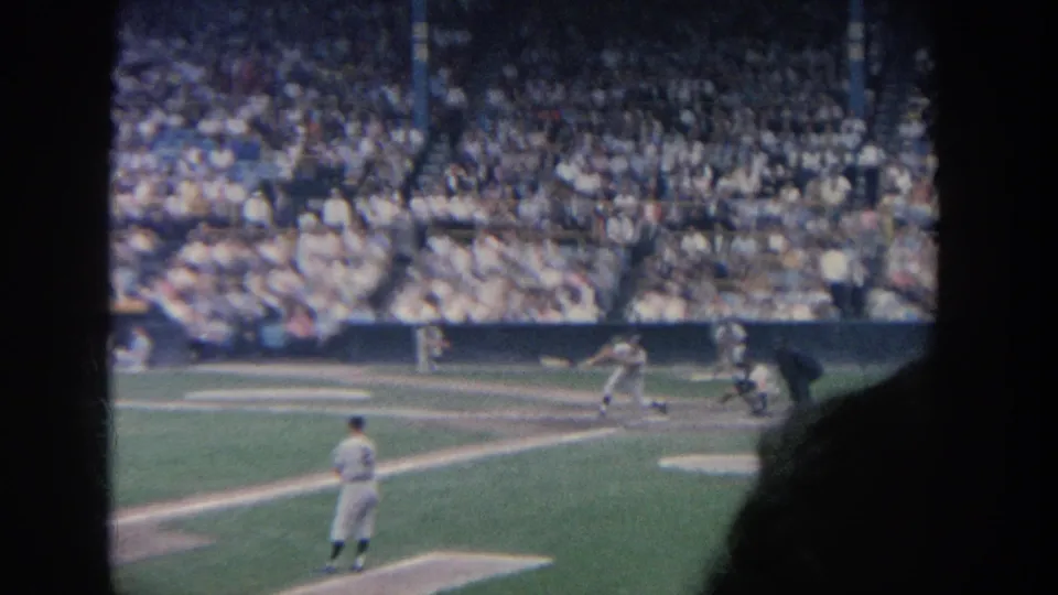 Distant 8mm view of a 1964 New York baseball at-bat, with home plate action partly framed by a spectator silhouette.