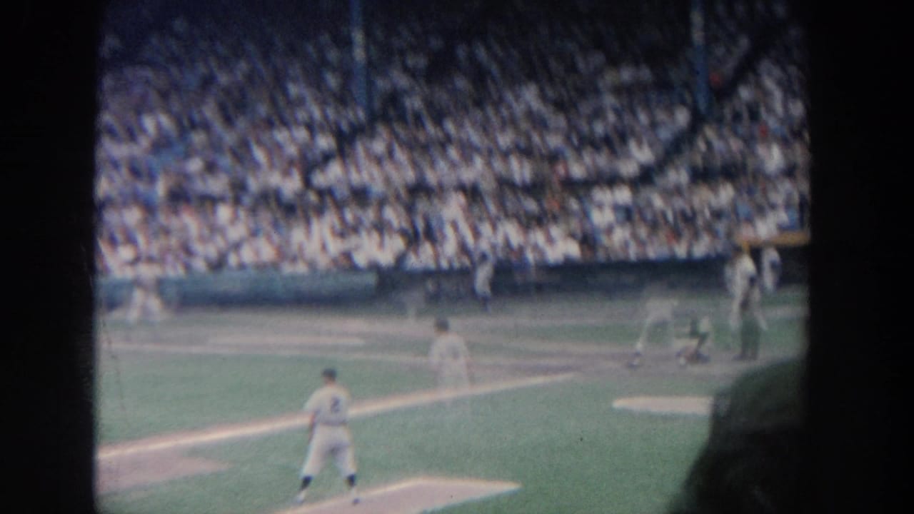 1964 New York home-movie view of a baseball game, with players in white uniforms on the field and a crowded grandstand in the background.