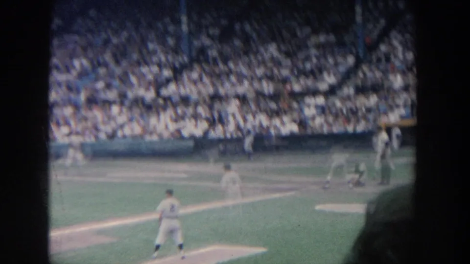 1964 New York home-movie view of a baseball game, with players in white uniforms on the field and a crowded grandstand in the background.