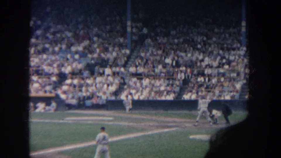 1964 New York baseball scene showing the batter at home plate, catcher and umpire nearby, a fielder in front, and dense stadium seating behind.