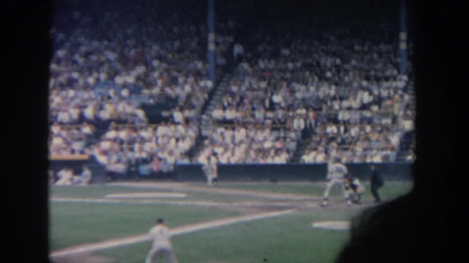 Spectator-shot 1964 New York baseball footage with home plate action, a foreground fielder, and a large crowd visible across the stadium seating.