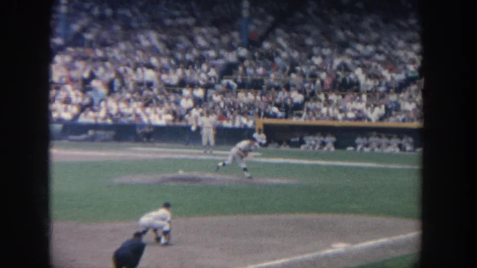 1964 New York home-movie frame with the pitcher in motion, batter at the plate, catcher low, and a full grandstand behind them.