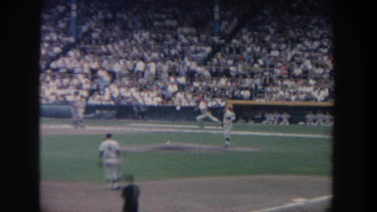 1964 New York 8mm frame of a baseball game, with players near the infield and a crowded grandstand in the background.