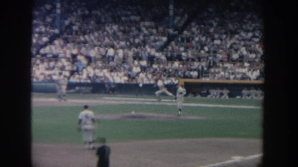1964 New York 8mm frame of a baseball game, with players near the infield and a crowded grandstand in the background.