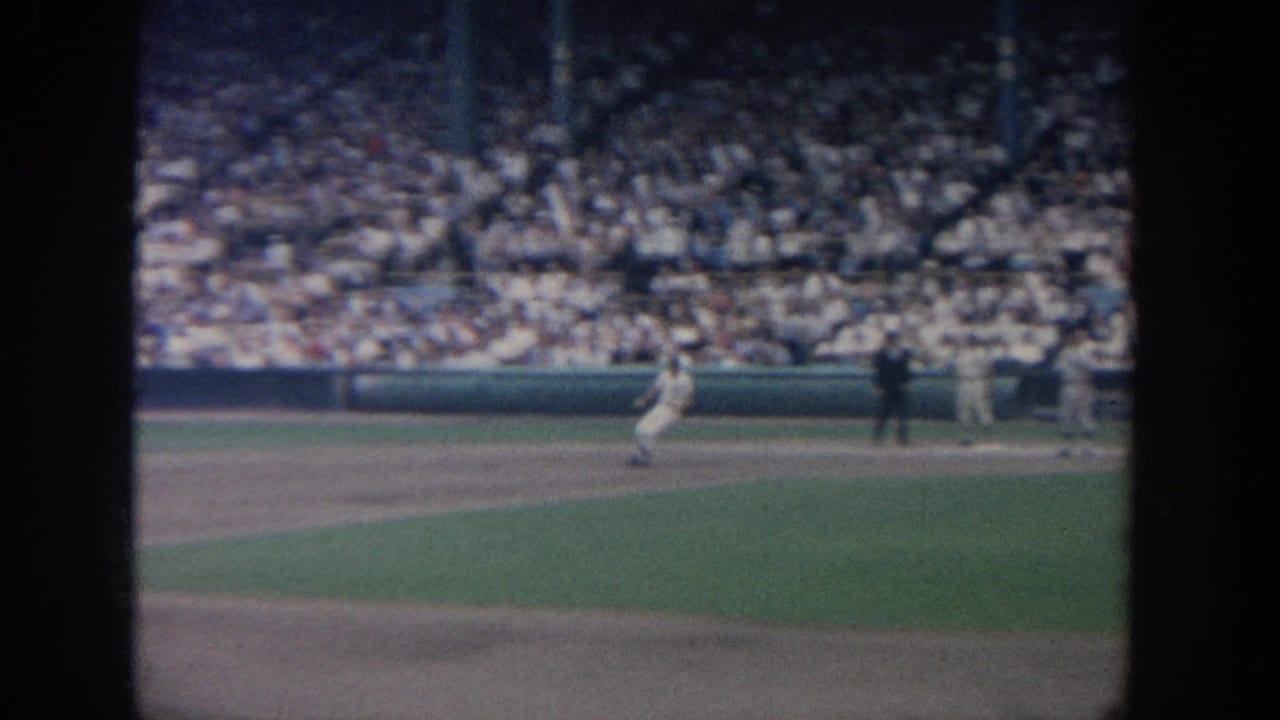 A batter or runner stands near the infield with spectators filling the stands.