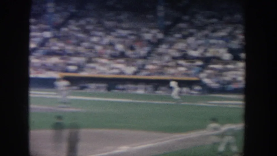 Soft 8mm frame from the 1964 New York baseball game, showing field action near a baseline with crowded seating beyond.