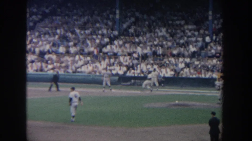 1964 New York 8mm baseball frame with players near the bases, an umpire in dark clothing, and packed stadium seating behind them.