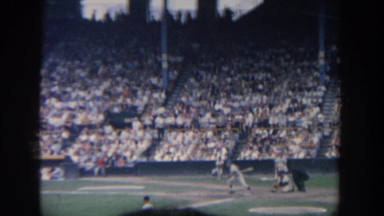 A baseball batter appears near home plate during a 1964 New York stadium game, with spectators filling the background.