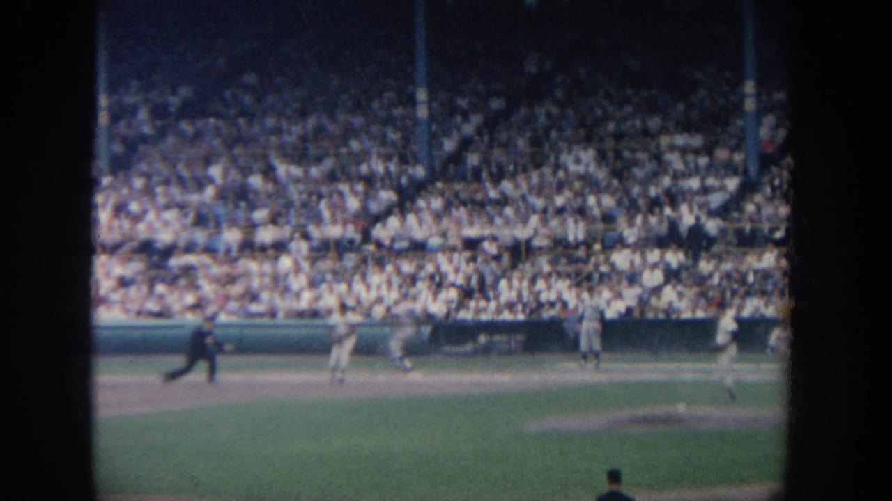 1964 New York 8mm frame showing a baseball game from the stands, with players on the infield and spectators filling the background.