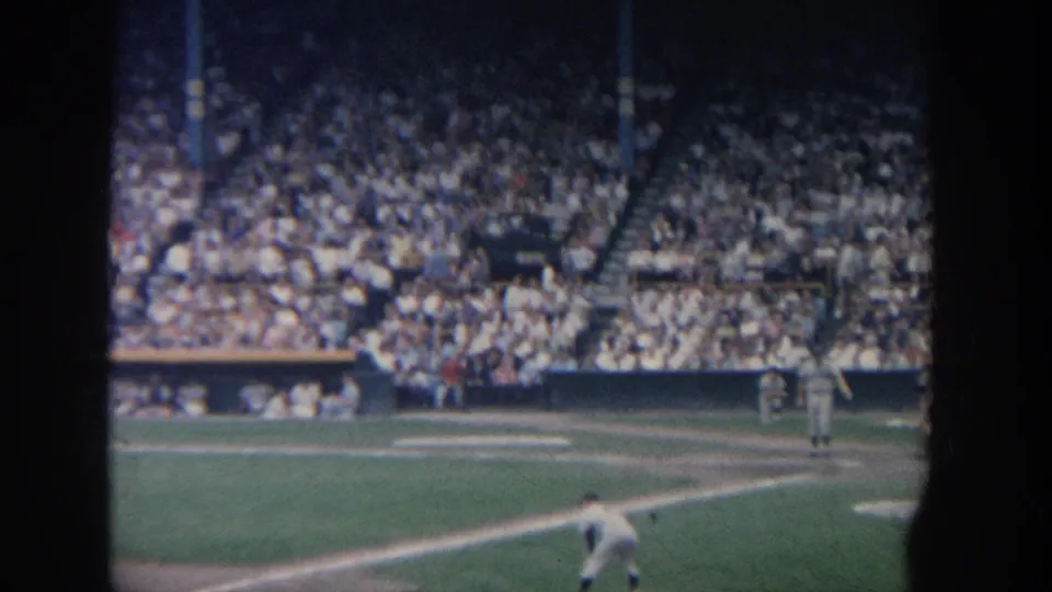 Wide 8mm view of a 1964 New York baseball game, with the mound in the foreground and a dense stadium crowd across the frame.