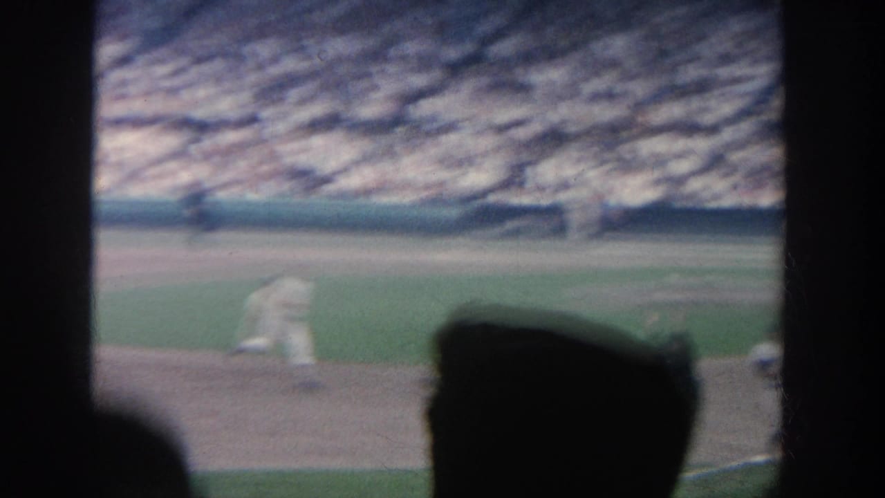 1964 New York 8mm view of a baseball game from the stands, partly obscured by dark foreground spectators and stadium framing.