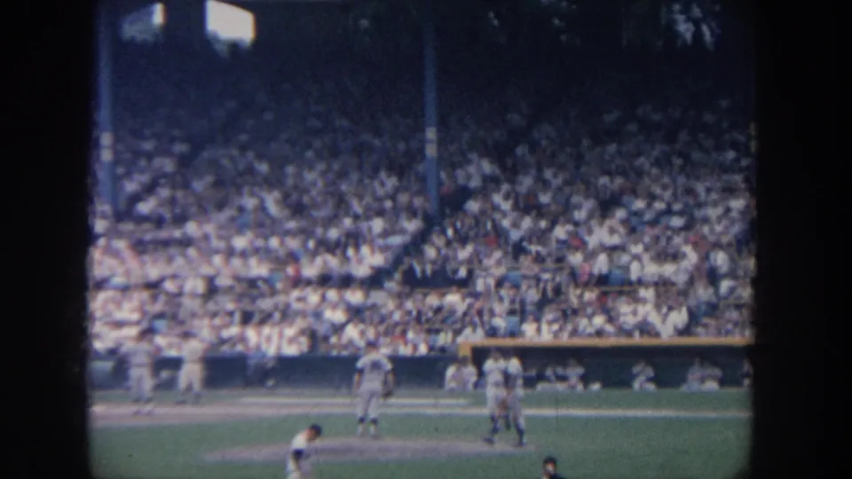 1964 New York home-movie frame of players near the infield, viewed from the stands with a crowded stadium behind.