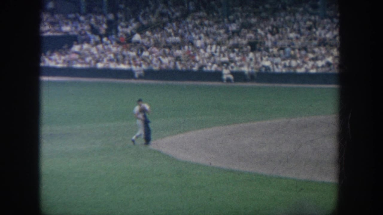 A baseball player walks near the dirt infield with crowded stadium seating behind him.
