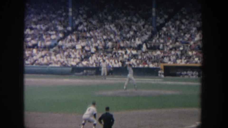 A distant 1964 New York baseball view shows a pitcher on the mound, foreground field figures, and crowded stands beyond.