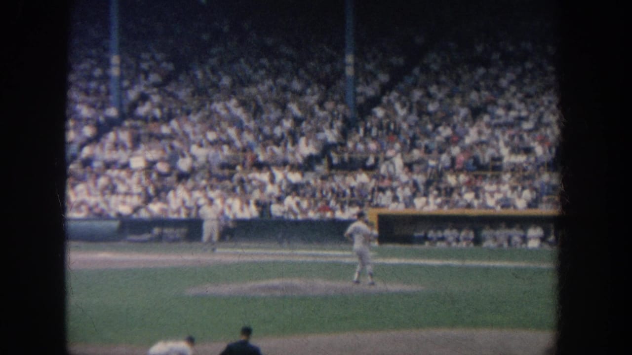 1964 New York 8mm frame showing a pitcher on the mound before a dense stadium crowd, seen from a spectator viewpoint.