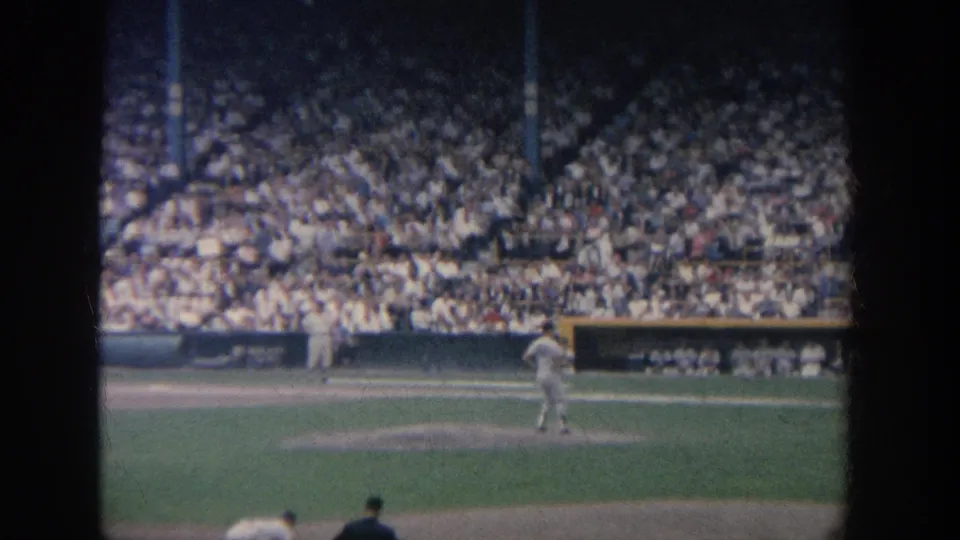 1964 New York 8mm frame showing a pitcher on the mound before a dense stadium crowd, seen from a spectator viewpoint.