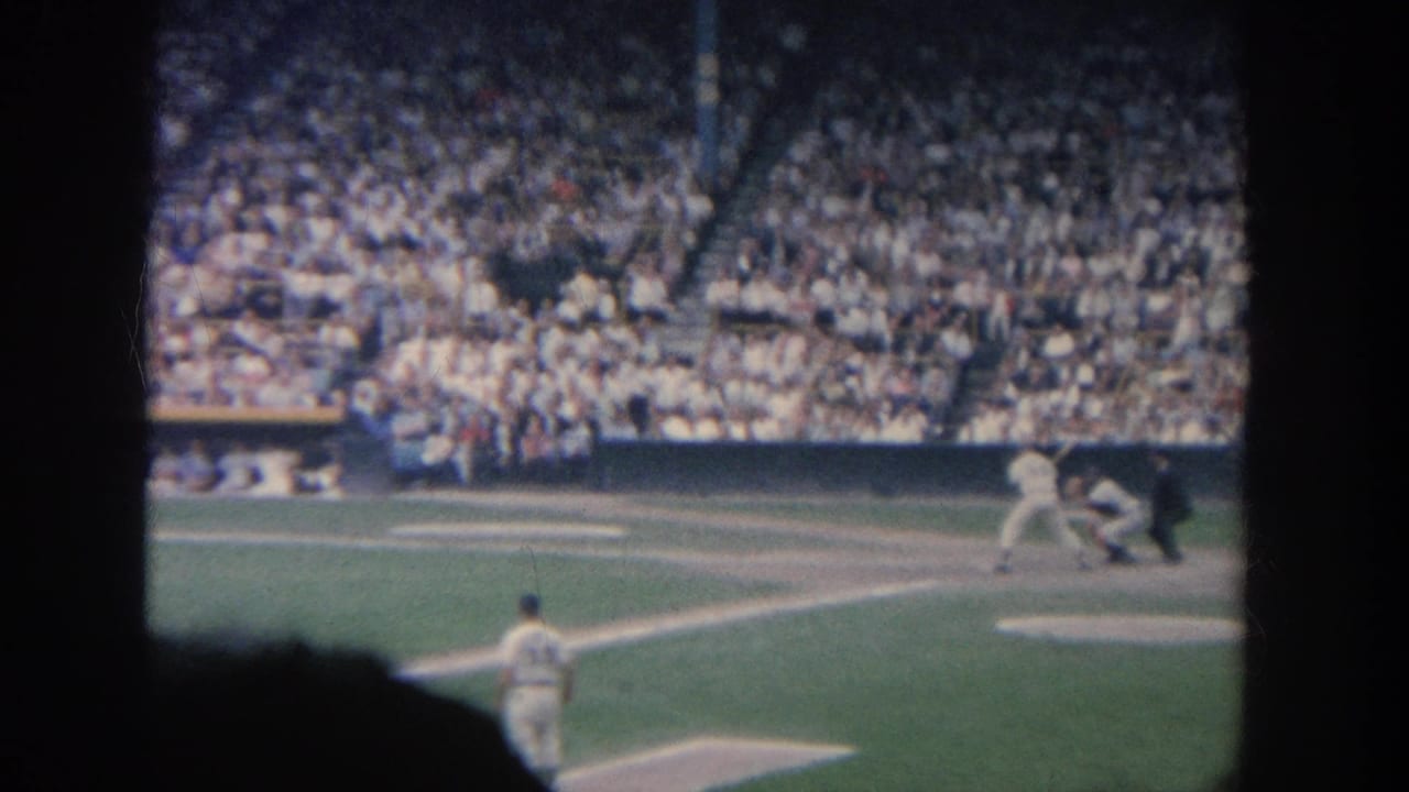 Batter, catcher, and umpire at home plate with players and crowd in the background.