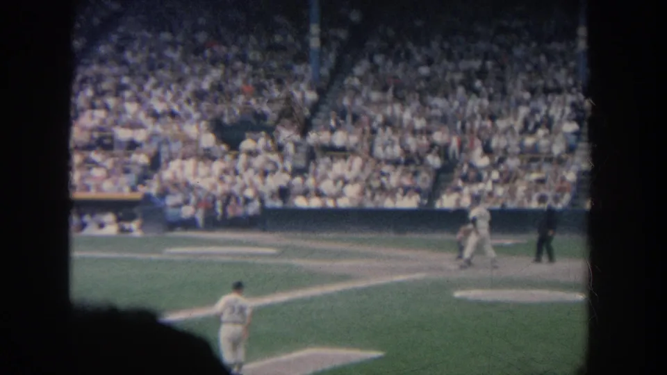 1964 New York 8mm frame with home plate action, an umpire, a foreground player, and packed grandstands in soft film color.