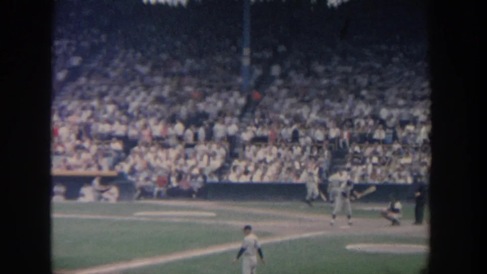 1964 New York home-movie frame showing baseball players on the field with the batter area and a dense stadium crowd behind them.