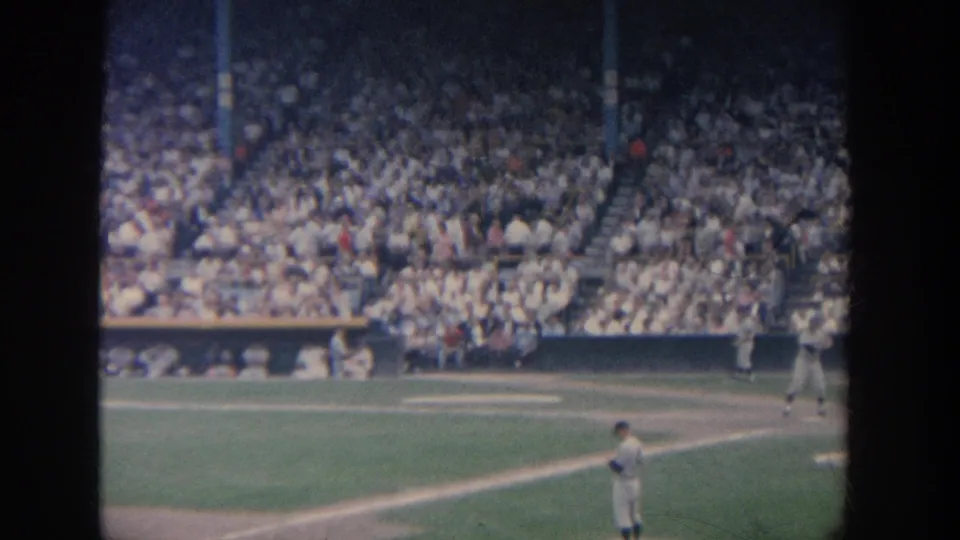1964 New York baseball frame with a player near the foreground, other players by the plate area, and spectators filling the stadium seats.