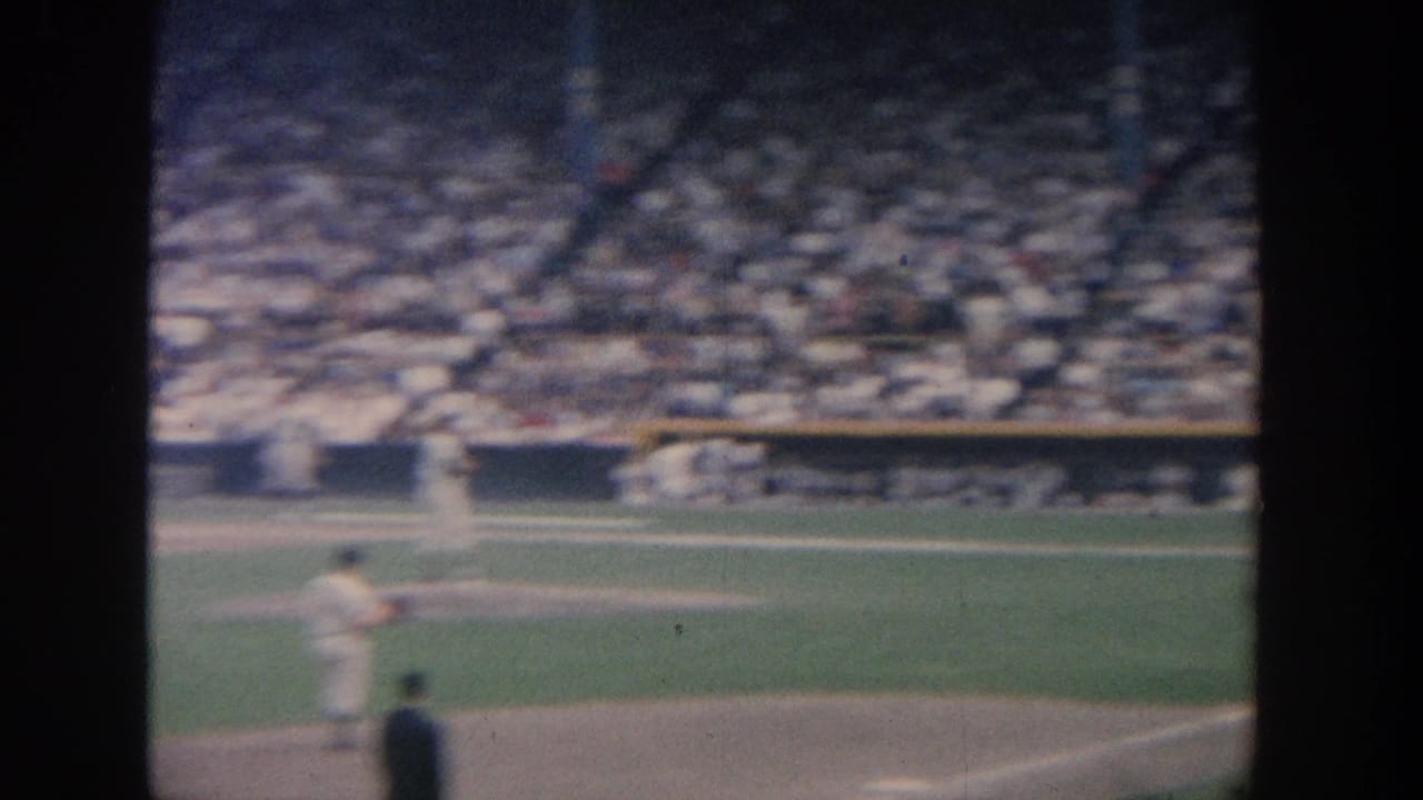 1964 New York 8mm home-movie view of a baseball pitcher on the mound, with home plate figures and crowded stadium seating in the background.