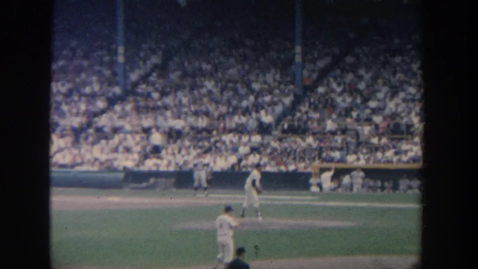 Wide 1964 New York stadium view showing a pitcher, batter, catcher, umpire, dugout area, and densely filled stands on soft 8mm film.