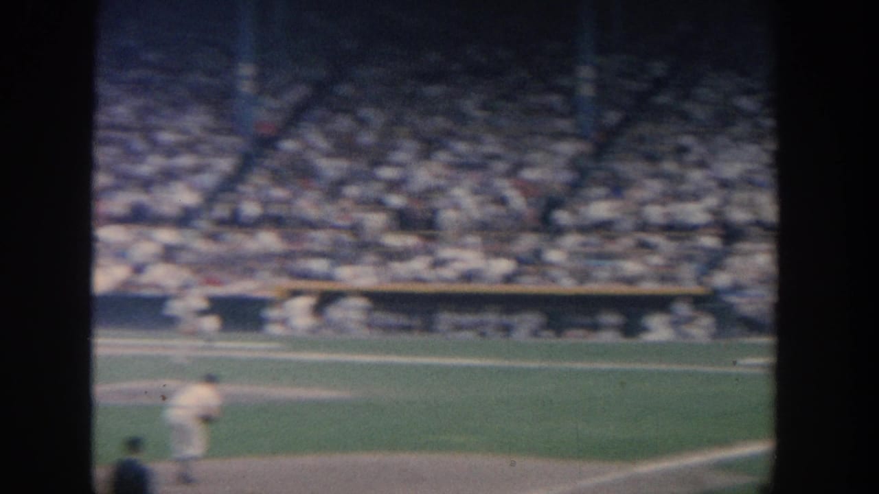 Grainy 1964 New York home-movie frame of baseball action, with a pitcher low in frame, an umpire nearby, and a full stadium behind the field.