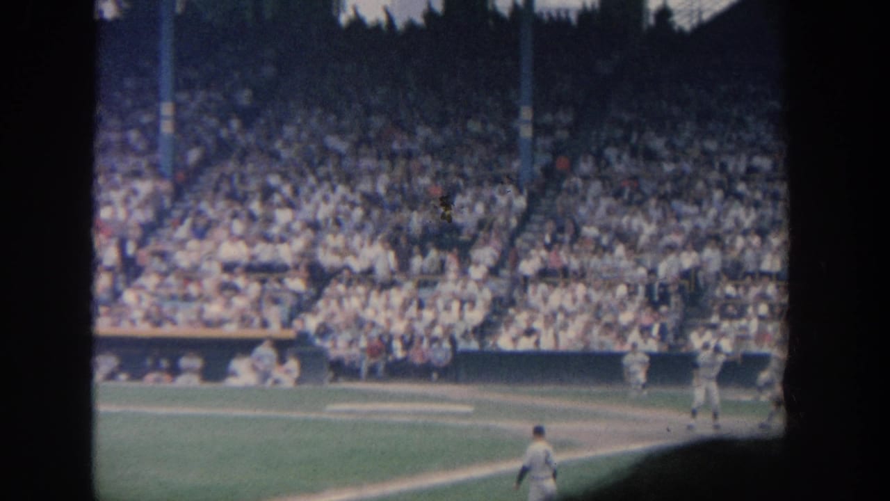1964 New York 8mm stadium frame showing the batter near home plate, pitcher on the field, catcher and umpire area, and a dense crowd in the stands.